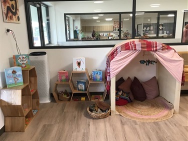 Indoor reading and play cubby sits next to a range honeycomb shaped shelves stacked with books, puzzles, puppets and other toys.