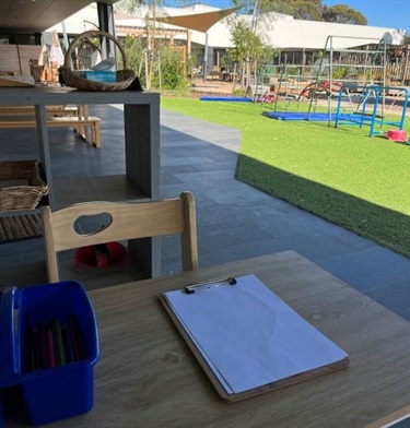 The table under the shaded veranda looks onto climbing equipment on the grass in the outdoor play area.